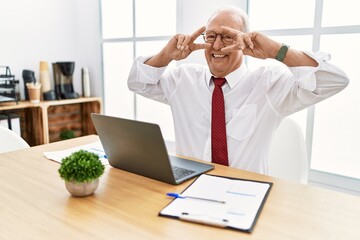 Senior man working at the office using computer laptop doing peace symbol with fingers over face, smiling cheerful showing victory