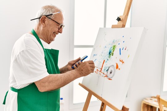 Senior grey-haired artist man smiling happy and using smartphone painting at art studio.