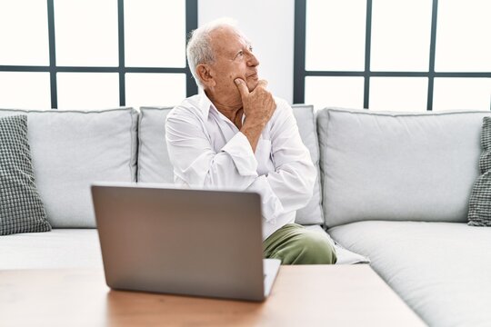 Senior man using laptop at home sitting on the sofa looking confident at the camera with smile with crossed arms and hand raised on chin. thinking positive.