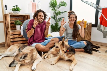 Young hispanic couple doing laundry with dogs gesturing finger crossed smiling with hope and eyes...
