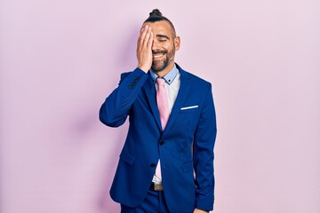 Young hispanic man wearing business suit and tie covering one eye with hand, confident smile on face and surprise emotion.