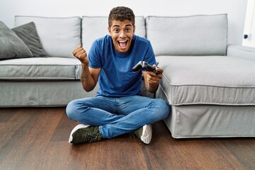 Young handsome hispanic man playing video game sitting by the sofa screaming proud, celebrating victory and success very excited with raised arms