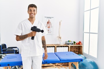 Young hispanic man working as physiotherapist holding massage gun at physiotherapy room