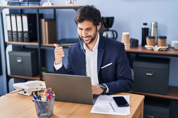 Handsome latin man working at the office using laptop pointing thumb up to the side smiling happy with open mouth