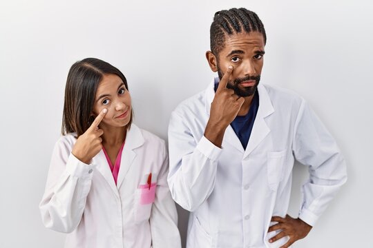 Young hispanic doctors standing over white background pointing to the eye watching you gesture, suspicious expression