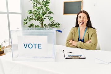 Young brunette woman sitting at election table with voting ballot sticking tongue out happy with...
