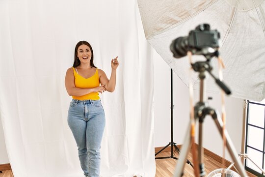 Young beautiful hispanic woman posing as model at photography studio with a big smile on face, pointing with hand and finger to the side looking at the camera.