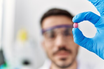 Naklejka premium Young hispanic man wearing scientist uniform holding pill at laboratory