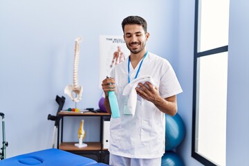 Young hispanic man wearing physiotherapist uniform holding disinfection sprayer at rehab clinic