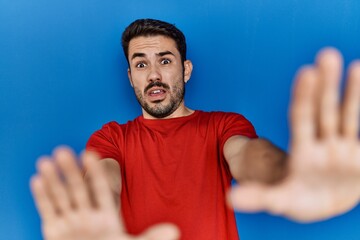 Young hispanic man with beard wearing red t shirt over blue background afraid and terrified with fear expression stop gesture with hands, shouting in shock. panic concept.