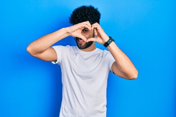 Young arab man with beard wearing casual white t shirt doing heart shape with hand and fingers smiling looking through sign