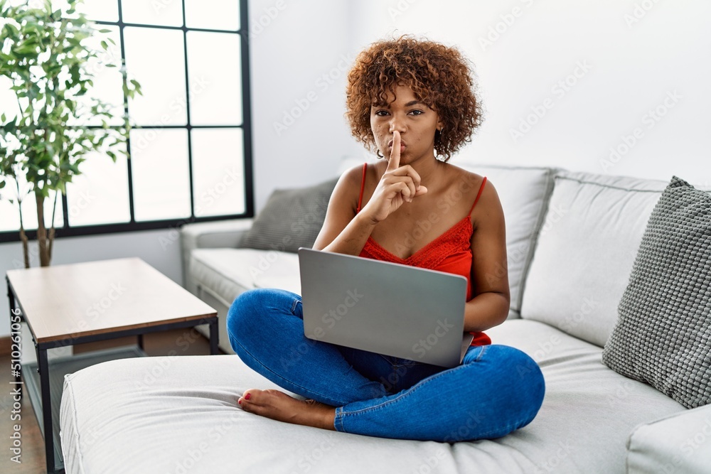 Sticker young african american woman sitting on the sofa at home using laptop asking to be quiet with finger