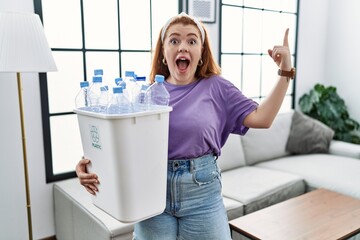 Young redhead woman holding recycling wastebasket with plastic bottles smiling amazed and surprised and pointing up with fingers and raised arms.