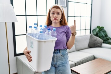 Young redhead woman holding recycling wastebasket with plastic bottles pointing up looking sad and...