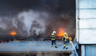 Firefighters at work. Several man are engaged in extinguishing fire. Firefighters stand on roof of burning building. Mans in firefighting uniform with his back to camera. Industrial building fire