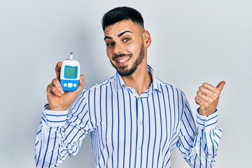 Young hispanic man with beard holding glucometer device pointing thumb up to the side smiling happy...