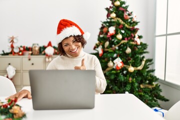 Fototapeta na wymiar Middle age hispanic woman using laptop sitting by christmas tree smiling happy and positive, thumb up doing excellent and approval sign