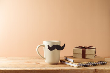 Father's day concept with coffee cup, mustache, notebook and gift box on wooden table over beige...