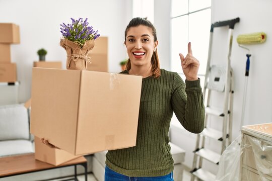 Young hispanic woman holding cardboard box at moving surprised with an idea or question pointing finger with happy face, number one