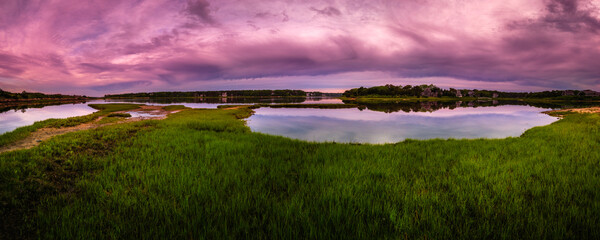 Rainbow seawater reflections, dramatic pink and purple cloudscape, green marshland wild plants, and tranquil coastal sunrise seascape on Cape Cod in June.