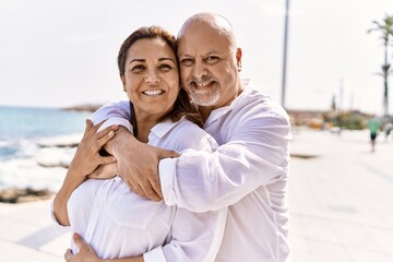 Obraz premium Middle age hispanic couple of husband and wife together walking by the beach on a sunny day. Hugging in love on vacation to the seaside.