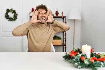 Young handsome man with beard sitting on the table by christmas decoration smiling cheerful playing...