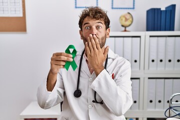 Young hispanic doctor man holding support green ribbon at clinic covering mouth with hand, shocked and afraid for mistake. surprised expression