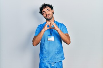 Young hispanic man wearing blue male nurse uniform smiling in love doing heart symbol shape with...