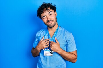 Young hispanic man wearing blue male nurse uniform winking looking at the camera with sexy...