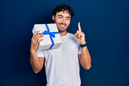 Young hispanic man holding gift smiling with an idea or question pointing finger with happy face, number one