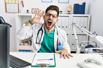 Fototapeta na wymiar Young man with beard wearing doctor uniform and stethoscope at the clinic shouting and screaming loud to side with hand on mouth. communication concept.