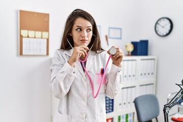 Young doctor woman wearing doctor coat holding stethoscope serious face thinking about question...