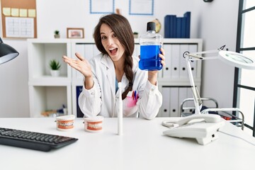 Young dentist woman holding mouthwash for fresh breath celebrating achievement with happy smile and winner expression with raised hand