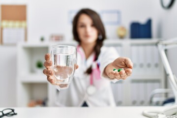 Young hispanic woman wearing doctor uniform holding pills and water glass at clinic