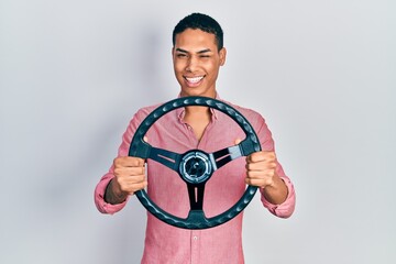 Young african american guy holding steering wheel winking looking at the camera with sexy expression, cheerful and happy face.