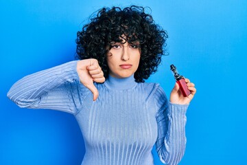 Young middle east woman holding electronic cigarette with angry face, negative sign showing dislike...