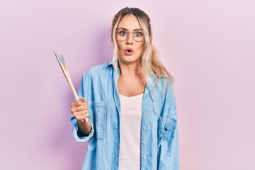 Beautiful young blonde woman holding paintbrushes scared and amazed with open mouth for surprise,...