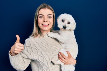 Beautiful caucasian woman hugging cute dog smiling happy and positive, thumb up doing excellent and approval sign