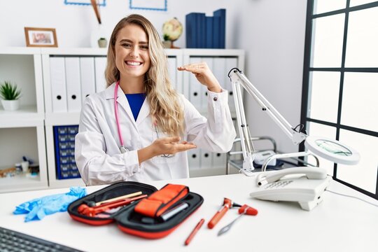 Young beautiful doctor woman with reflex hammer and medical instruments gesturing with hands showing big and large size sign, measure symbol. smiling looking at the camera. measuring concept.