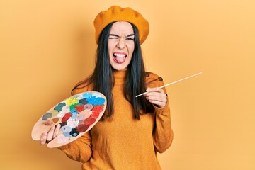 Young brunette woman holding paintbrush and palette wearing beret sticking tongue out happy with funny expression.