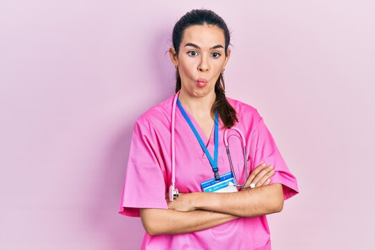 Young brunette woman wearing doctor uniform and stethoscope standing with arms crossed making fish face with mouth and squinting eyes, crazy and comical.