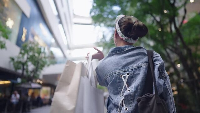 Back behind of Asian happy tourist shopper enjoys lifestyle walking in the shopping mall alone in daytime. Cheerful fashionable girl carrying paper bags leaving the shop after spending on sunny day