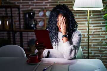 Middle age hispanic woman using touchpad sitting on the table at night yawning tired covering half...