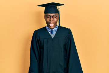 Young african american man wearing graduation cap and ceremony robe sticking tongue out happy with...