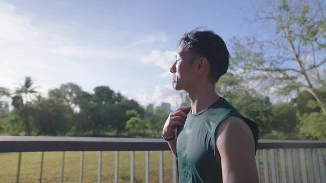 Handsome Asian man with his backpack walking along the park walking bridge get ready to do some exercise at the park after work, workout motivation, heading to workout, side view slow motion shot