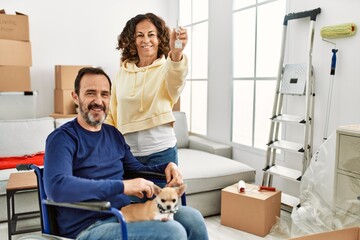 Middle age hispanic couple smiling happy. Man sitting on wheelchair with dog on his legs and woman holding key of new home.