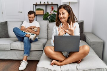 Obraz premium Hispanic middle age couple at home, woman using laptop disgusted expression, displeased and fearful doing disgust face because aversion reaction. with hands raised