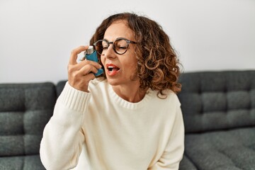 Mature hispanic woman using asthma inhaler sitting on the sofa at home - obrazy, fototapety, plakaty