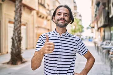 Young hispanic man with beard outdoors at the city doing happy thumbs up gesture with hand. approving expression looking at the camera showing success.