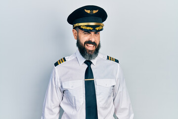 Young hispanic man wearing airplane pilot uniform winking looking at the camera with sexy expression, cheerful and happy face.